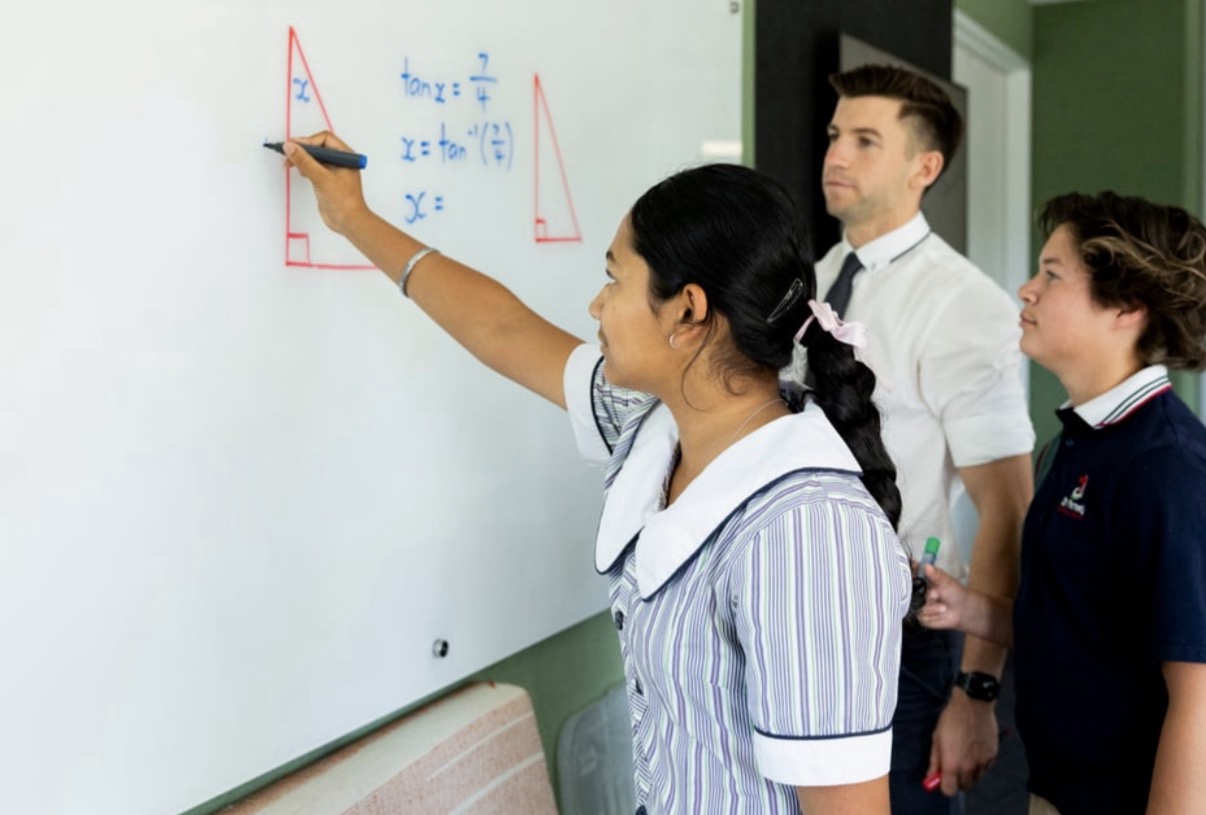 A student working through mathematics at a whiteboard alongside a teacher and another student